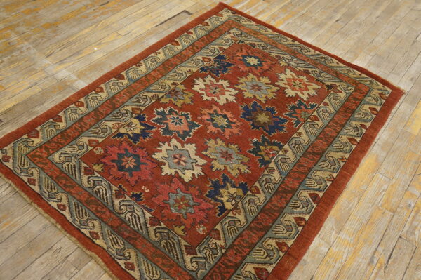 Reddish-orange rug featuring blue, cream, and green geometric motifs with multiple patterned borders on wood planks.