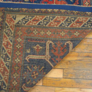 A corner of a navy, red, and tan geometric rug is folded back on distressed wood plank flooring.