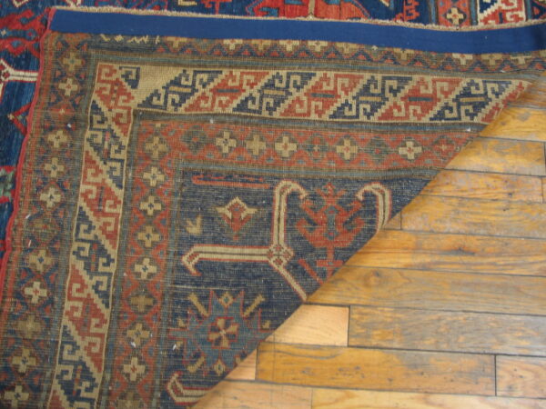 A corner of a navy, red, and tan geometric rug is folded back on distressed wood plank flooring.