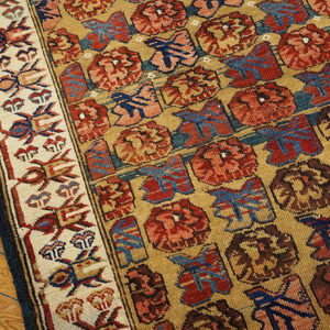Low-pile rug corner showing red and blue repeating motifs on a gold field, resting on plank wood flooring.