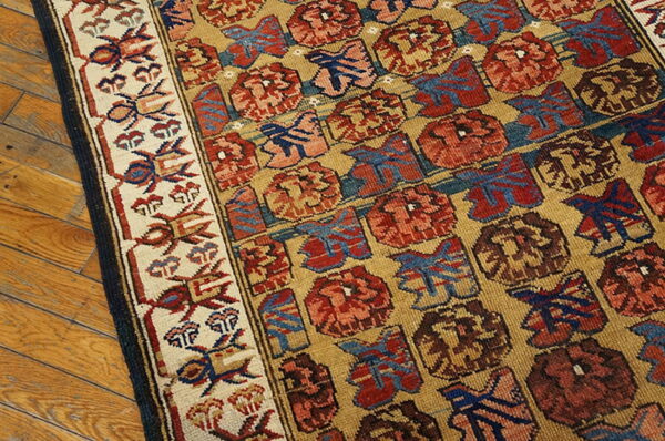 Low-pile rug corner showing red and blue repeating motifs on a gold field, resting on plank wood flooring.
