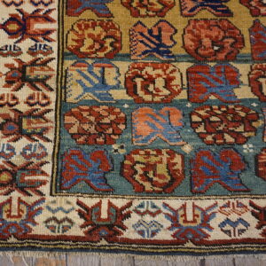 Geometric rug with rows of red and blue motifs on a teal and gold background, bordered in ivory.