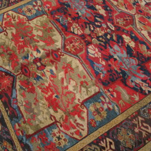 Close view of a richly patterned rug featuring red, khaki, and blue traditional panels and borders on a wooden floor.