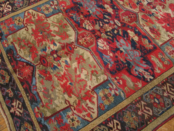 Close view of a richly patterned rug featuring red, khaki, and blue traditional panels and borders on a wooden floor.