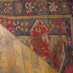 Detail of a patterned red and blue rug folded back, resting on worn, light brown wood planks.