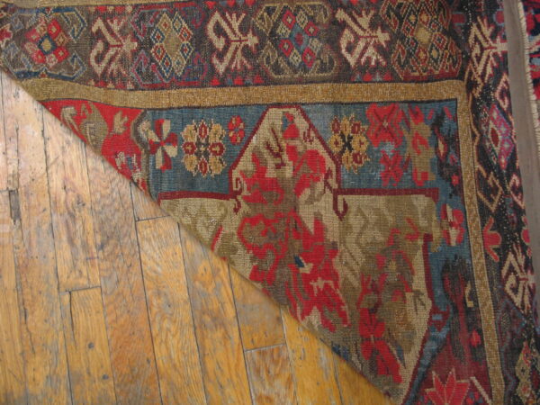 Detail of a patterned red and blue rug folded back, resting on worn, light brown wood planks.