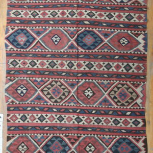 Flatwoven rug with red, navy blue, and cream geometric diamond patterns over light wood planks.