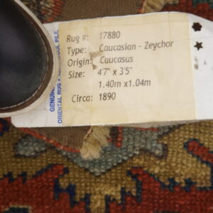 A geometric pattern rug in deep red, olive, and gold, obscured by an identifying tag and a dark shoe.