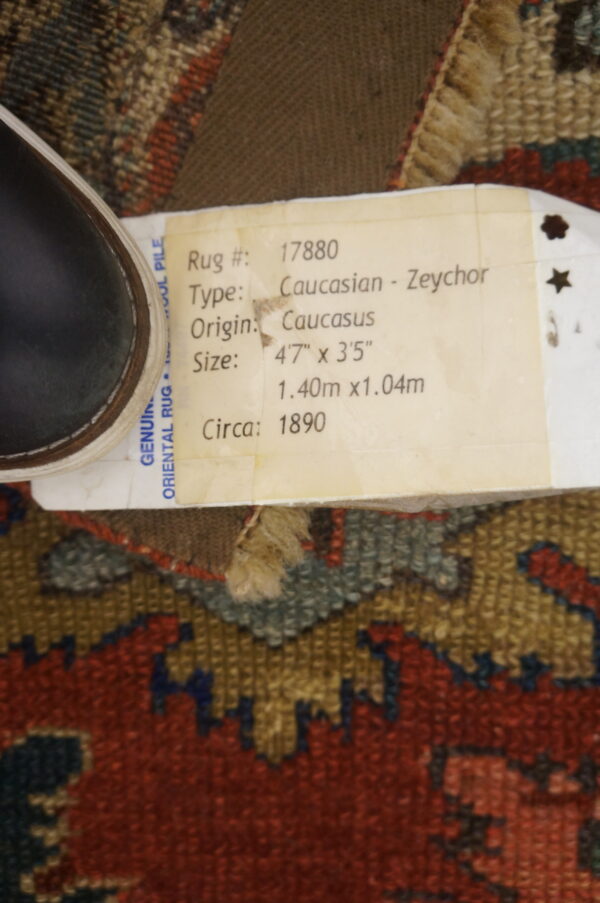 A geometric pattern rug in deep red, olive, and gold, obscured by an identifying tag and a dark shoe.