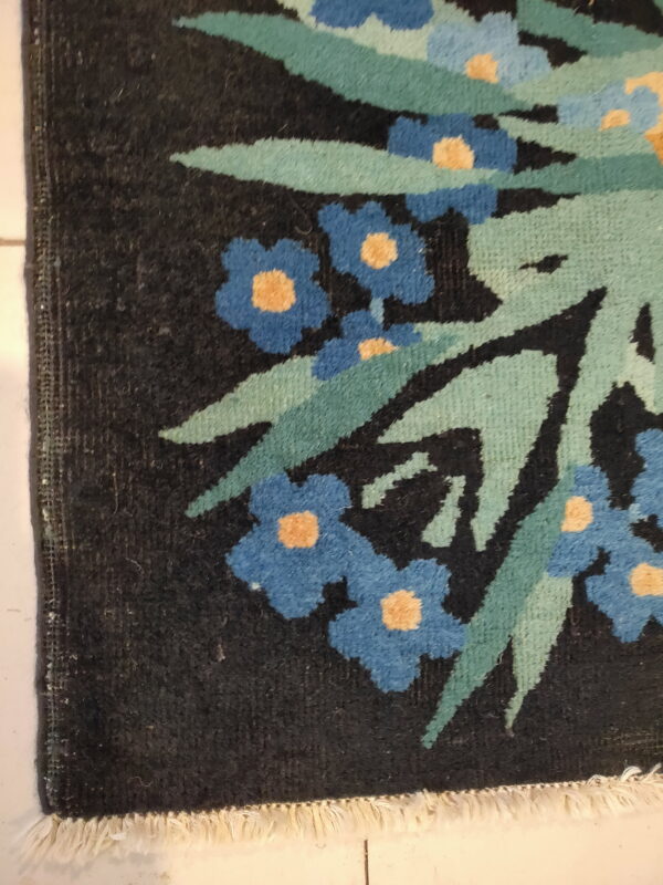 Dark floral rug detail showing blue and yellow flowers, green foliage, and white fringe on light tile.