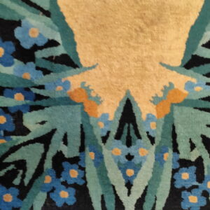 Close-up detail of a textured rug featuring blue and orange flowers, teal leaves, black patterns, and a tan field.