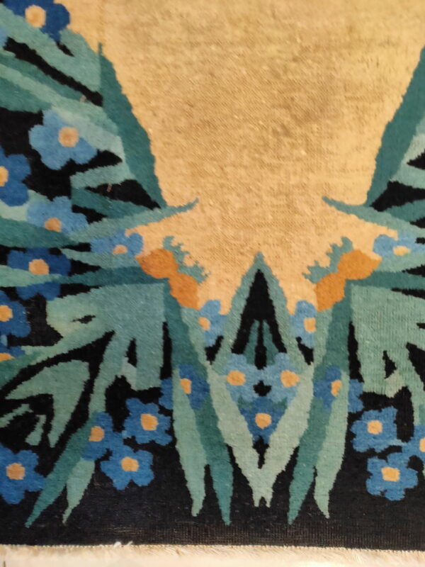 Close-up detail of a textured rug featuring blue and orange flowers, teal leaves, black patterns, and a tan field.