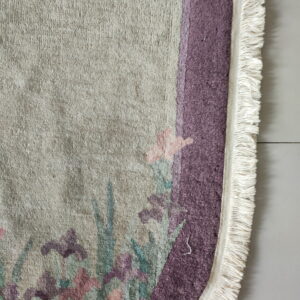 Textured beige rug section showing dusty purple and pink floral patterns and white woven fringe on light flooring.