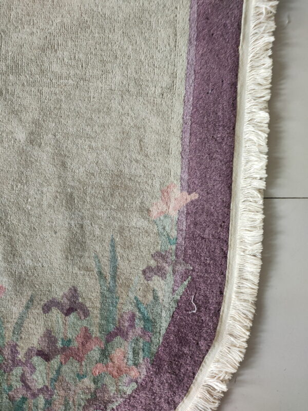 Textured beige rug section showing dusty purple and pink floral patterns and white woven fringe on light flooring.