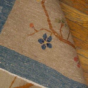 A tan rug, featuring a blue border and delicate floral pattern, is partially curled on a wood plank floor.