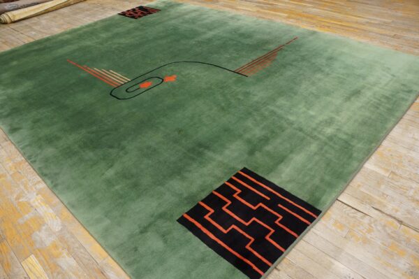 Low-pile green rug with abstract black and orange geometric patterns lies on worn wooden flooring.
