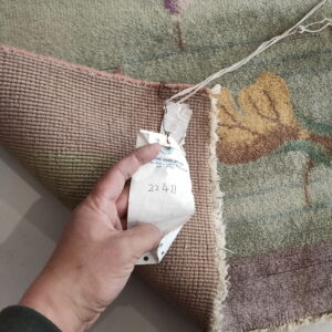Low-pile green rug with purple and gold patterns; a hand holds a tag against the pink woven backing.