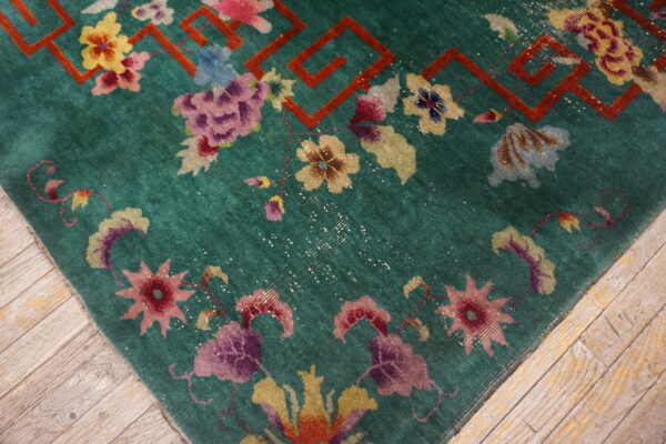 Teal rug with vibrant floral designs and a red geometric border rests on distressed wooden floorboards.