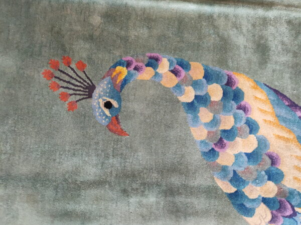 Detail of a rug showing a multicolor peacock design against a subtly textured sage green background.