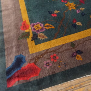 Teal rug with multicolored floral pattern, featuring gold and taupe borders, rests on worn wood flooring.