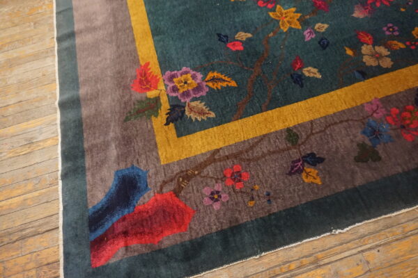 Teal rug with multicolored floral pattern, featuring gold and taupe borders, rests on worn wood flooring.