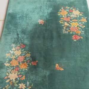 Velvety green rug with vibrant floral and butterfly patterns, framed by fringe on white tile.