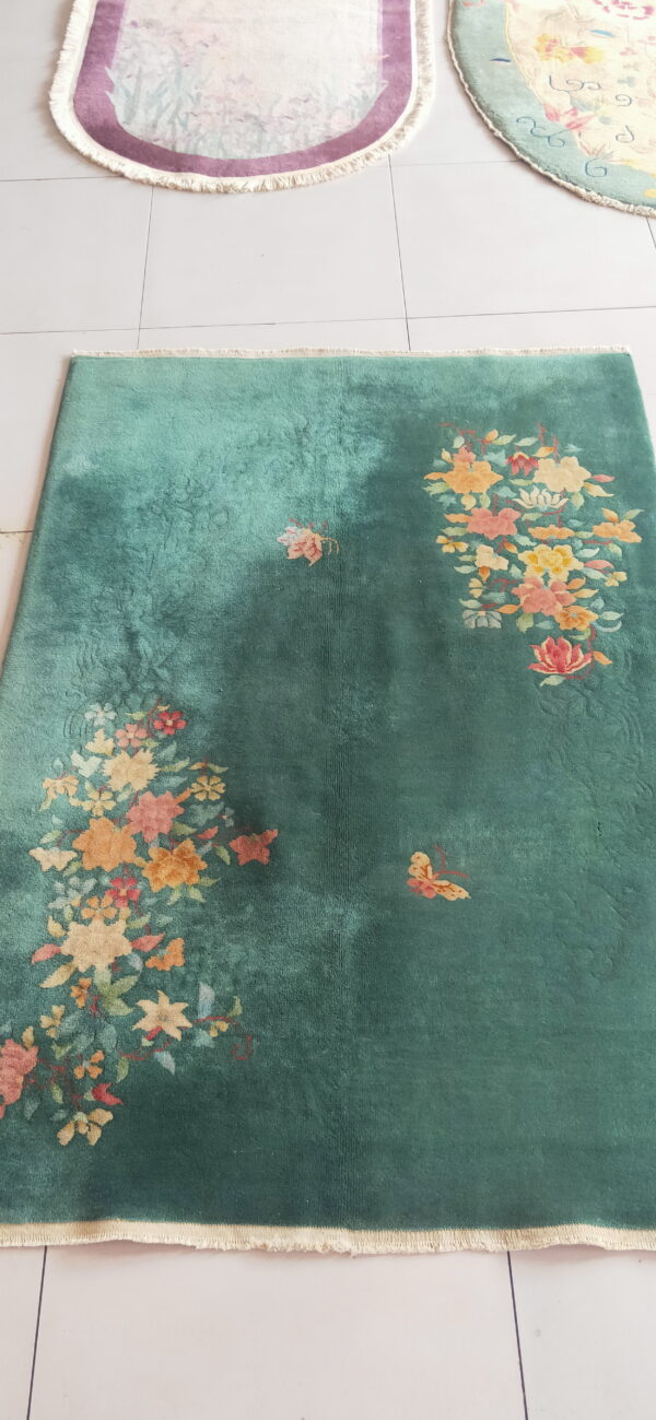 Velvety green rug with vibrant floral and butterfly patterns, framed by fringe on white tile.