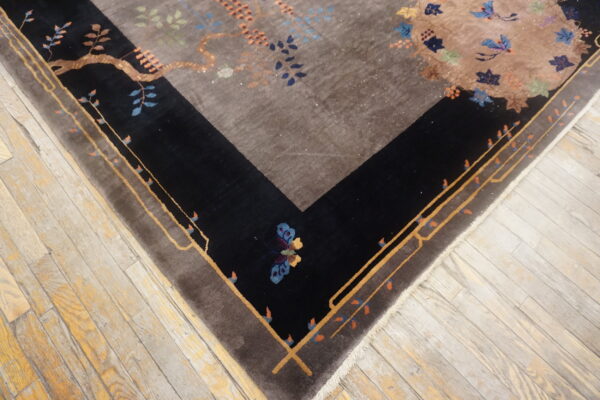 High-pile rug with a taupe center and black, stylized floral border resting on worn wood planks.