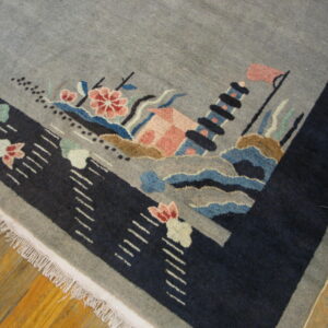 Gray rug corner with a navy border and fringe, displaying a multicolored scenic design on wood floors.