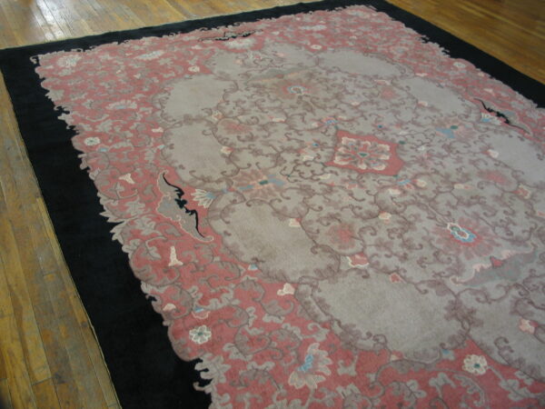 Ornate gray and dusty rose low-pile rug with a wide black border rests on light brown hardwood flooring.