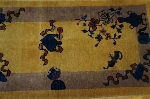 Gold rug with a wide taupe border featuring stylized blue, red, and brown figures and floral patterns.