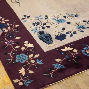 Beige rug corner featuring a deep burgundy border, blue floral motifs, and a central decorative vase on wood flooring.