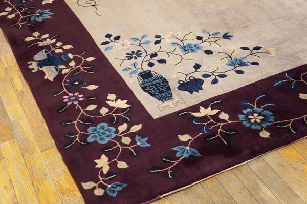 Beige rug corner featuring a deep burgundy border, blue floral motifs, and a central decorative vase on wood flooring.
