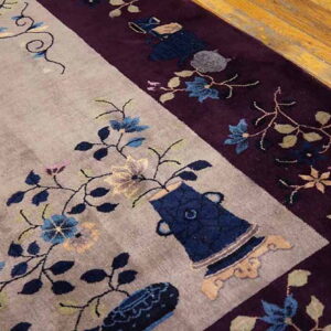 Carved rug detail showing a beige field, plum border, and blue floral and vase patterns on wood flooring.