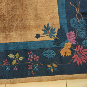 Textured tan rug corner with a deep blue botanical border and multicolor flowers on light tile flooring.