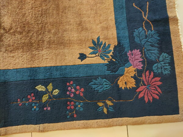 Textured tan rug corner with a deep blue botanical border and multicolor flowers on light tile flooring.
