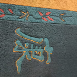 High-pile charcoal rug detail featuring a teal and orange carved symbol bordered by a red and green vine pattern.