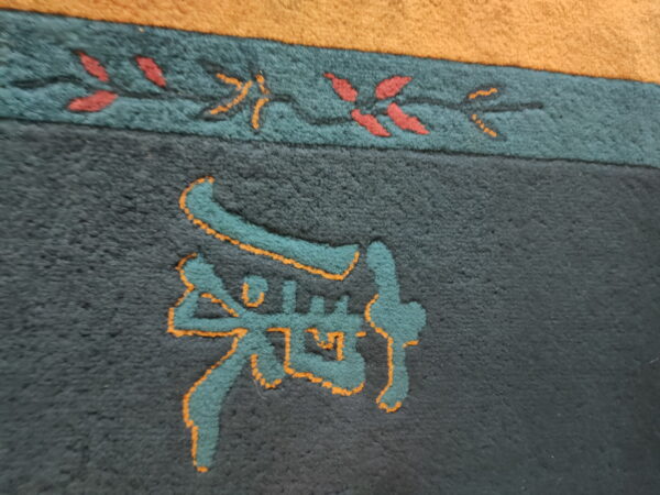 High-pile charcoal rug detail featuring a teal and orange carved symbol bordered by a red and green vine pattern.