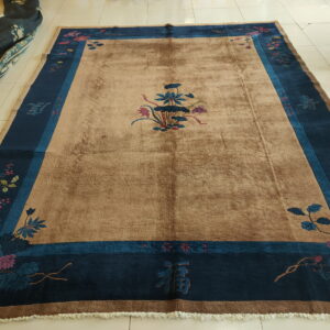 A tan rug with a plush pile features a central flower motif and a wide navy and teal floral border on tile.