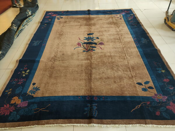 A tan rug with a plush pile features a central flower motif and a wide navy and teal floral border on tile.