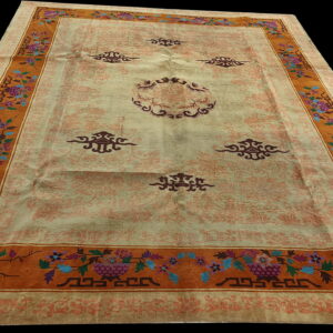 Light beige rug with maroon central motifs and a wide orange border featuring colorful floral and fret patterns.