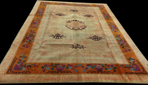 Light beige rug with maroon central motifs and a wide orange border featuring colorful floral and fret patterns.