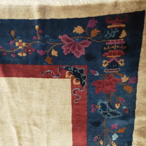 Corner of a cream rug featuring a deep blue border with ornate multicolored floral and vine motifs on white tiles.