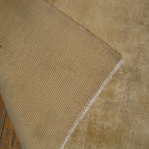 Tan, low-pile rug corner folded back, revealing a dense ribbed backing on wooden planks.