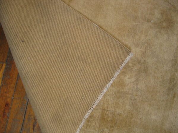 Tan, low-pile rug corner folded back, revealing a dense ribbed backing on wooden planks.