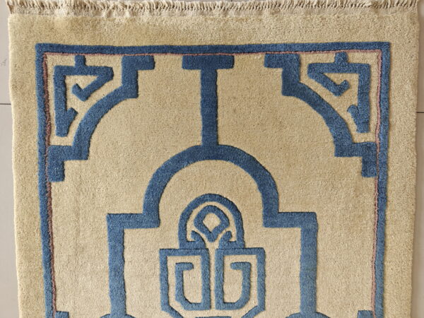 Textured beige rug with bold blue geometric design, thin pink outline, and visible fringes.