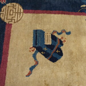 Close-up of a rug with a tan field, navy and pink border, and blue patterned motif.
