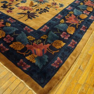 Plush gold rug corner featuring a dark blue border with red, pink, and teal floral designs on wood floors.