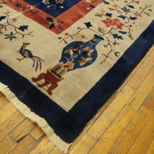 Close-up of a tan rug with floral, bird, and vase motifs and a wide navy border on wood planks.