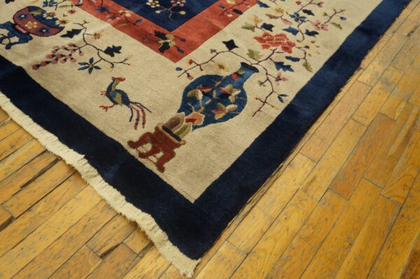 Close-up of a tan rug with floral, bird, and vase motifs and a wide navy border on wood planks.